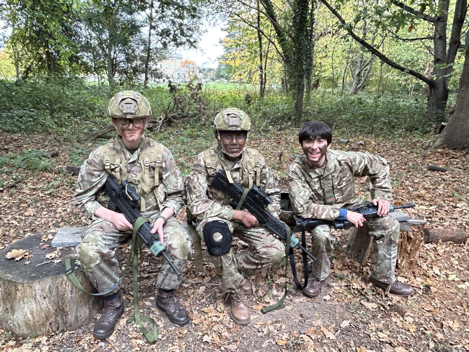 Three young people in camouflage uniforms, helmets, and face paint are sitting and resting in a wooded area. They are holding training rifles, indicating they are taking a break during a field exercise.