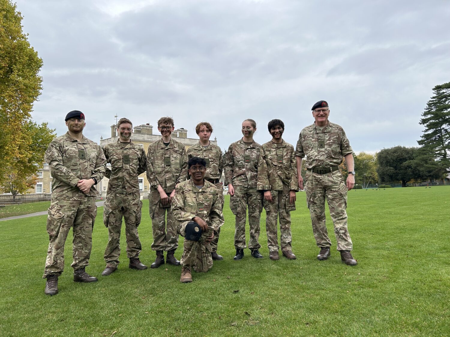 A group of eight military or cadet personnel, dressed in camouflage and berets, posing for a portrait on a green lawn in front of a stately building. They appear to be an organized training unit.