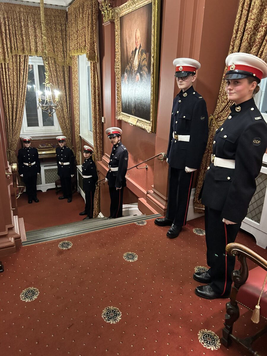 Five young people in military-style uniforms (black and red with white caps and belts) are positioned on a grand staircase and landing. Two are standing at attention on the landing in the foreground, facing slightly towards the right. Three others are standing at attention further back on the stairs leading down. The surroundings are ornate and historical, featuring red carpet with decorative gold medallions, heavy gold-colored drapes framing a window, and a large, dark-framed portrait hanging on the wall above the stairs. The space has a very formal and traditional atmosphere.