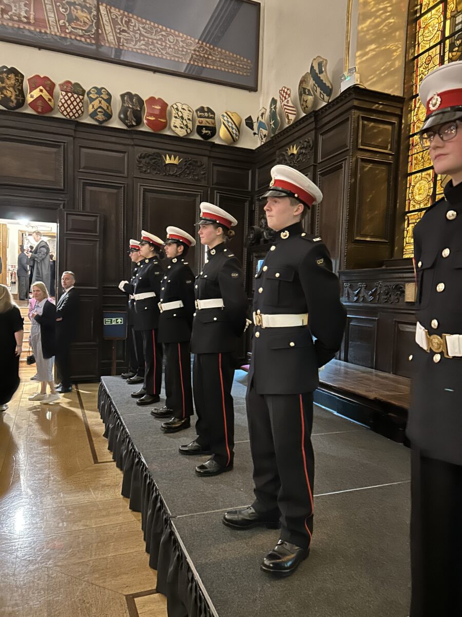 A group of six young people, all dressed in black and red military-style uniforms with white belts and white-topped caps, are standing in formation on a low, dark platform. They appear to be acting as a guard of honor or display team. The setting is a grand, wood-paneled hall, possibly a livery company or college hall, with a high ceiling. Above the dark wood paneling, there is a frieze of colorful heraldic shields or crests. In the background on the left, a few adults in civilian clothes are visible near a doorway, observing the group. The lighting is warm, highlighting the polished wooden floor and the uniforms.