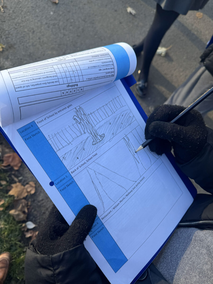 A close-up, overhead view of a person wearing black gloves and using a pencil to draw a field sketch on a data collection sheet attached to a clipboard. The sketch, labeled "Location 4: Front of School by entrance gates," depicts a fenced area with leafless trees. The clipboard also holds a rolled-up checklist, showing sections for rating "Buildings" and "Land use" attributes.