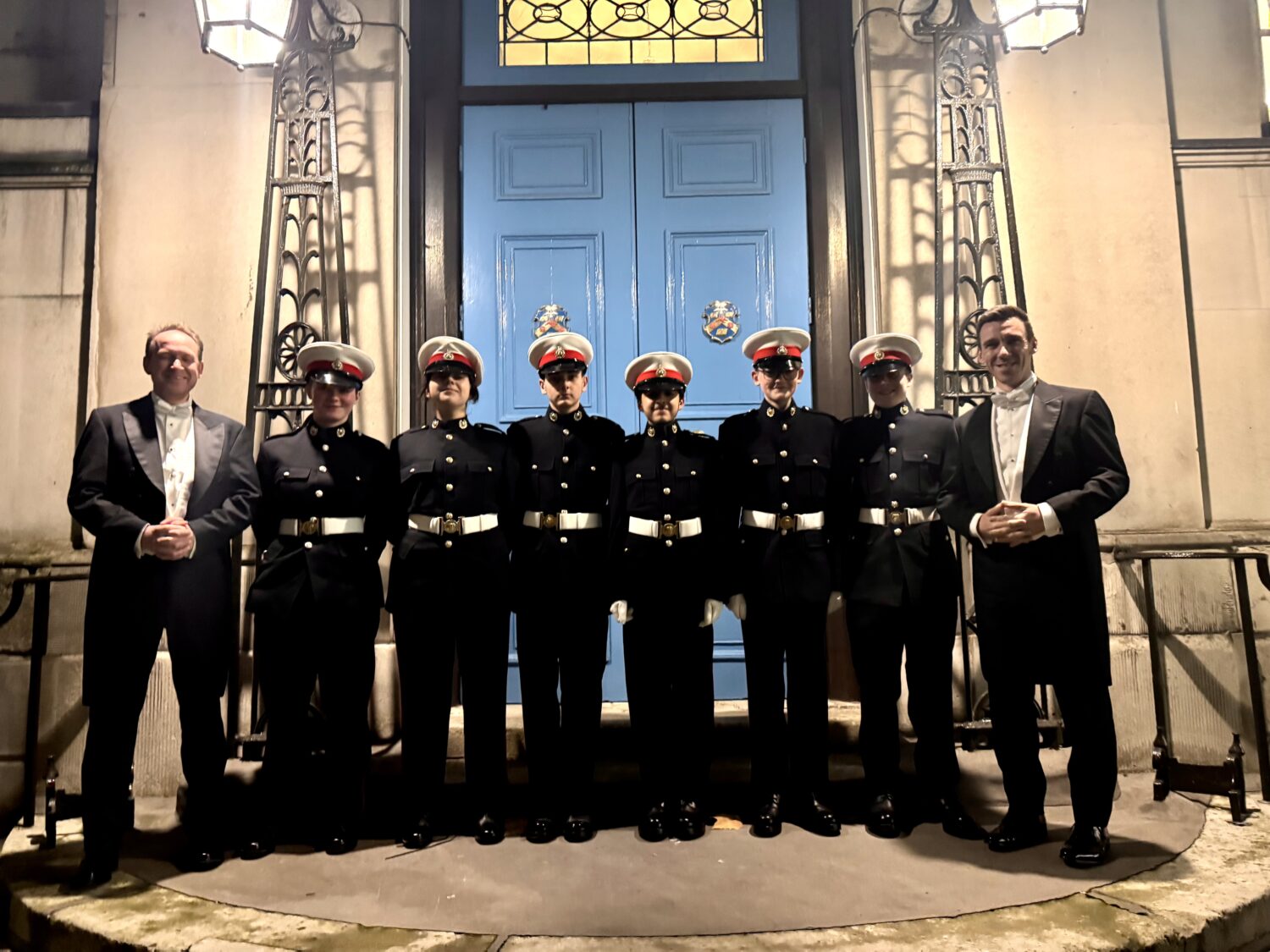 A line of eight people is posed at night in front of a grand building entrance with light blue double doors. Six of the people in the center are young people wearing the same dark military-style uniform with white belts and caps. On either end of the line are two adults dressed in formal evening wear: black tuxedo jackets, bow ties, and white shirts. Above the group, the entrance is flanked by ornate wrought-iron light fixtures, which illuminate the blue doors and the people standing below. The atmosphere is formal, suggesting a ceremonial or special evening event.