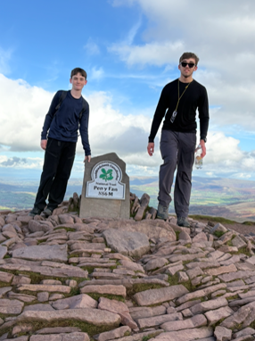 Two hikers stand next to the Pen y Fan summit sign (886 M) in Wales, celebrating their successful climb on a sunny day.