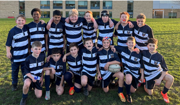 A group of twelve boys, likely a youth sports team, is posing for a photo outdoors on a grassy field in front of a modern school building. They are all wearing matching rugby kit, consisting of dark blue and white horizontal striped jerseys, dark blue shorts, and dark socks. The boys are arranged in two rows—a standing back row and a front row kneeling or crouching. Several of the boys have their arms around each other's shoulders, and one boy in the front row is holding a Gilbert rugby ball. Many of the boys have grass or mud stains on their legs, suggesting they have just finished playing a match. The field is bathed in sunlight, and a long, low school building with large windows forms the background.