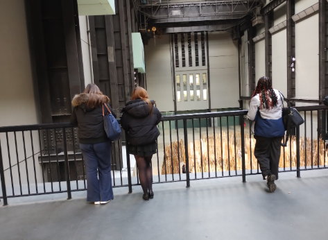 Three people, viewed from behind, standing at a black railing on an upper level of a large industrial interior space, looking down into the vast Turbine Hall of the Tate Modern. The hall's high walls are visible, and the floor below is filled with an art installation that appears to involve light or reflective golden-brown materials.
