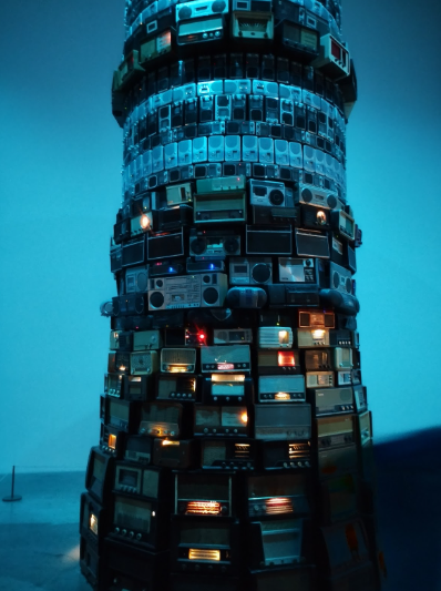 A tall, cylindrical contemporary sculpture constructed entirely out of vintage and antique radios, boomboxes, and stereo components. The artwork is dramatically lit with a cool blue light from above, making the overall structure appear blue, while some individual radios glow with warm, yellow, and red lights from their dials and power indicators.