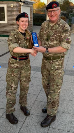 A photo of a cadet in camouflage uniform and a beret smiling while shaking hands with an older man in a matching camouflage uniform and glasses. The man is presenting her with a blue-hued award or trophy, which they are both holding. They are standing outdoors on a paved area near a building with horizontal wooden siding.
