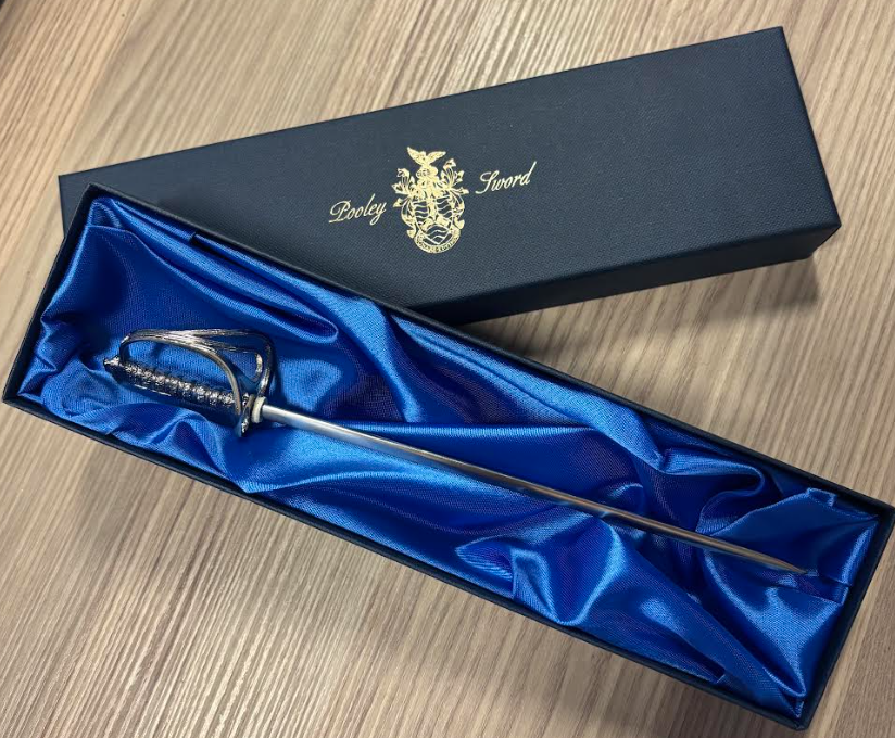 A silver miniature officer's sword, resembling a ceremonial basket-hilted sword, resting in a long, narrow, dark blue box lined with bright royal blue satin. The lid of the box is open and features the gold text "Pooley Sword" with a coat of arms logo above the sword.