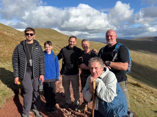 A group of six hikers is posing on a mountain trail, taking a break during their climb up the grassy slopes.
