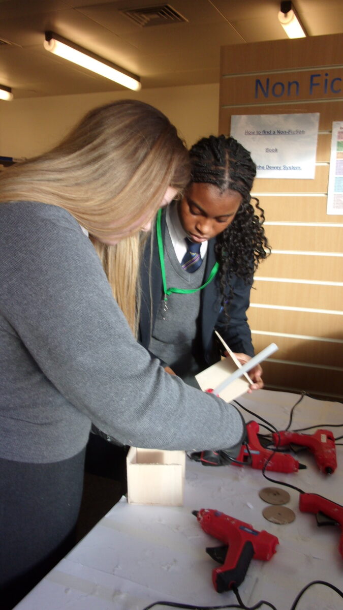 Two female students, one with long blonde hair and one with dark, braided hair, are leaning over a table working on a craft project. They are handling small pieces of wood or cardboard. Several red glue guns are placed on the table next to their project. A sign in the background reads "Non Fic[tion] How to find a Non-Fiction Book using the Dewey System."