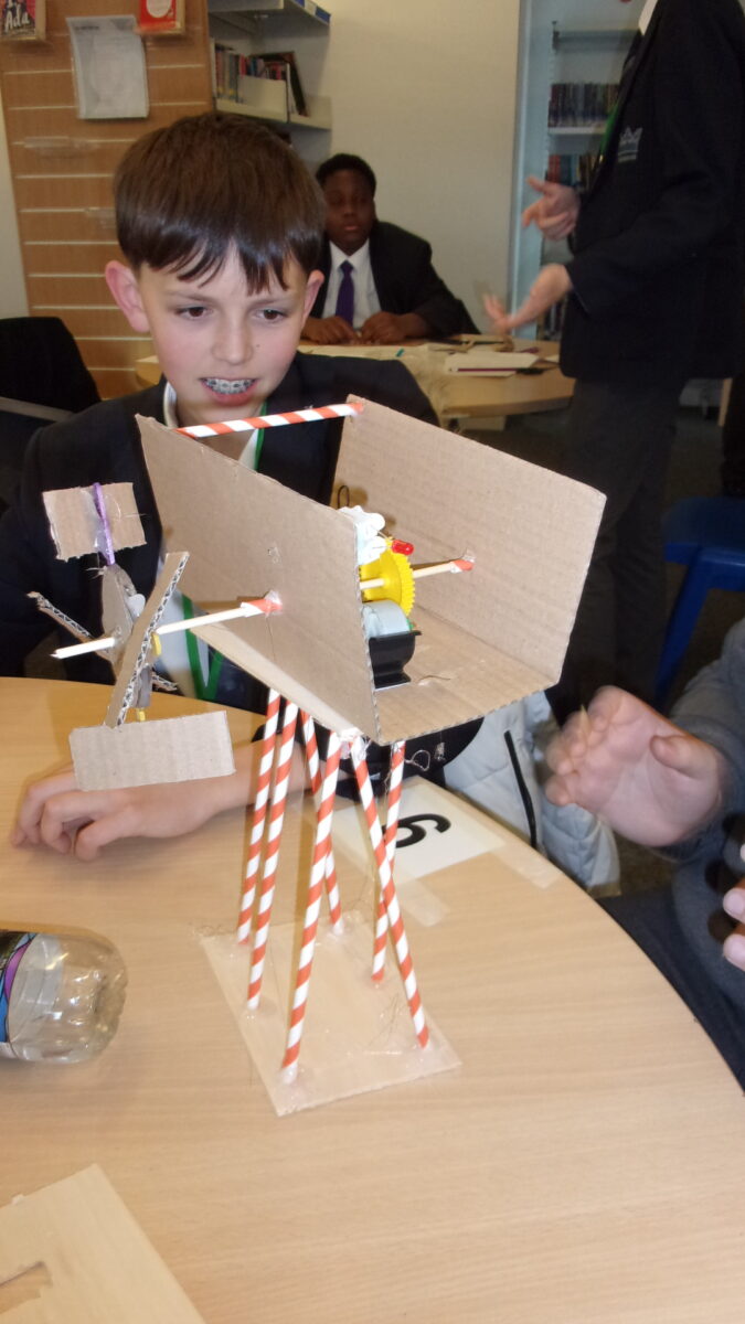 A male student in a dark blazer and tie, wearing braces, looks intently at a model project on a wooden table. The project, which appears to be a wind or water power generator, is constructed from cardboard, straws, and a small yellow plastic gear. Another student's hands are visible on the table.