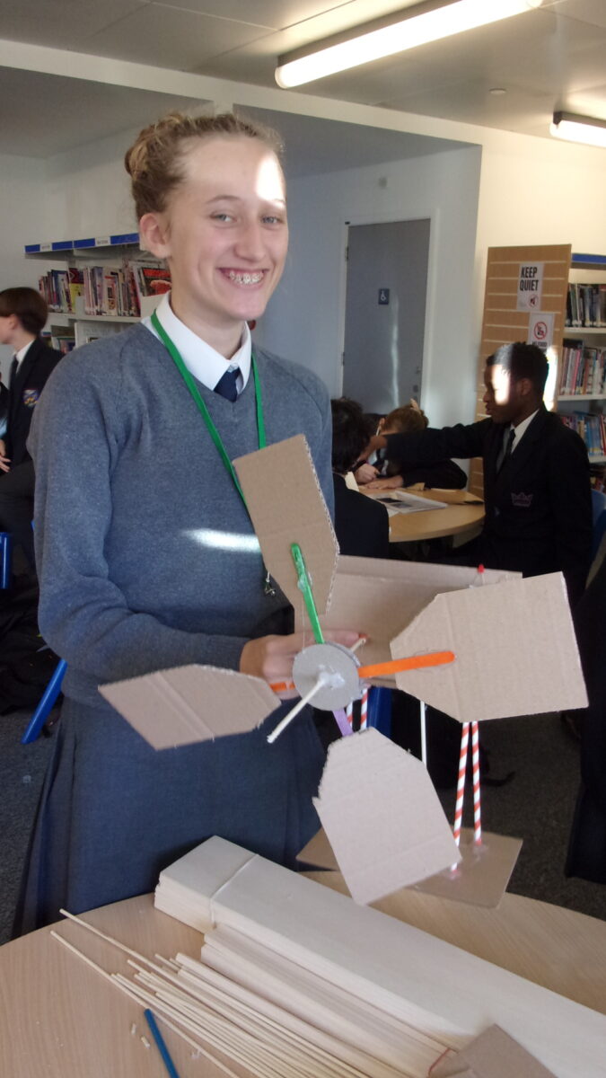 A smiling female student in a gray sweater and blue skirt stands in a classroom holding a model wind turbine or watermill made of cardboard and plastic straws. The model features three blades and a structure supported by red and white striped straws. She is wearing braces and a green lanyard. Other students are visible in the background, which includes bookshelves.