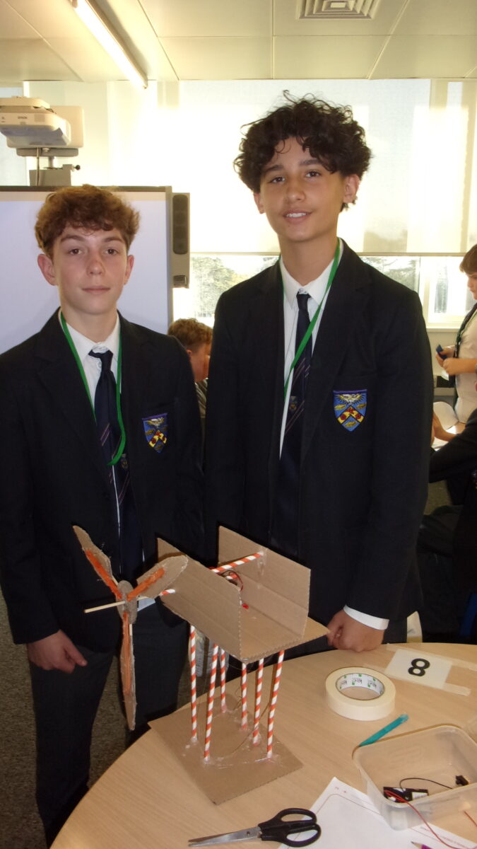 Two male students in matching dark blazers, ties, and school badges stand side-by-side behind a project model. The model is made of cardboard and straws and is supported by several red and white striped straws. The boys are smiling, and a whiteboard and projector screen are visible in the background.