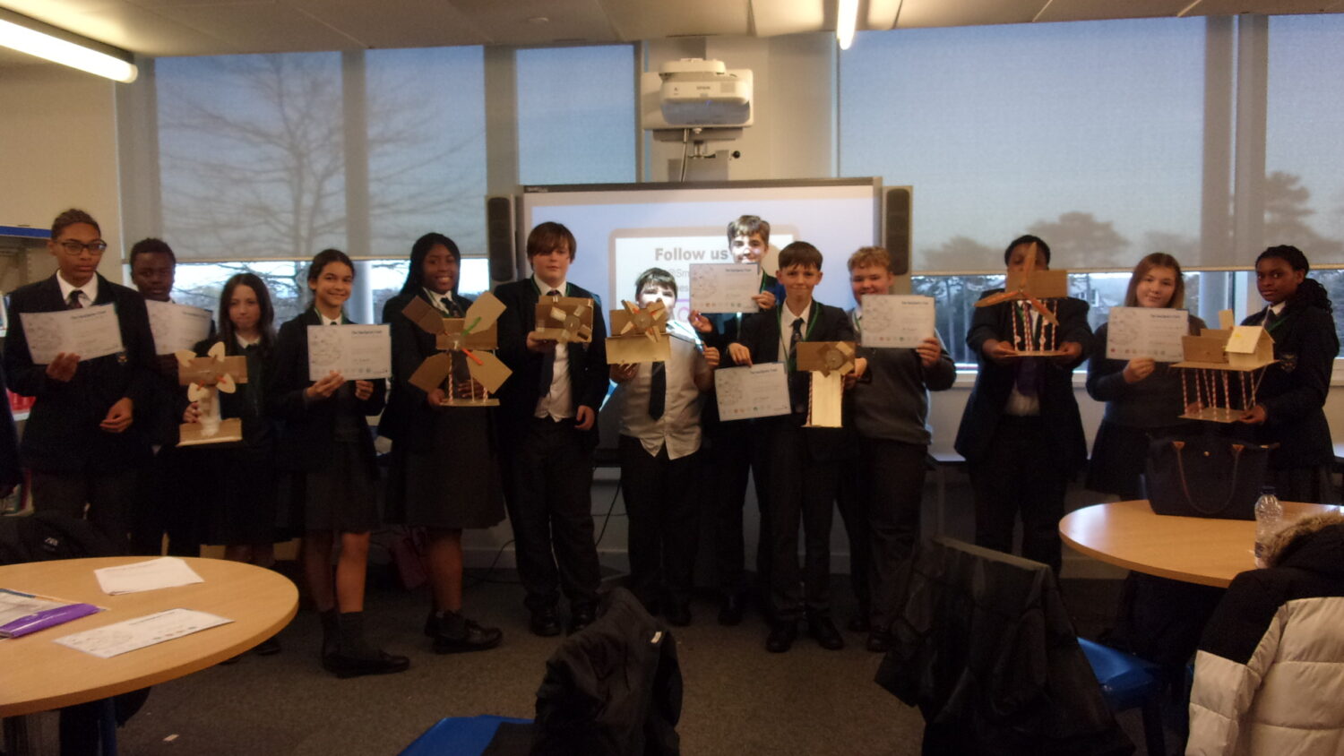 A group of approximately ten students, dressed in school uniform, stand together in a classroom, holding up their completed energy challenge models and certificates. They are posing in front of a projector screen, celebrating the conclusion of the project.