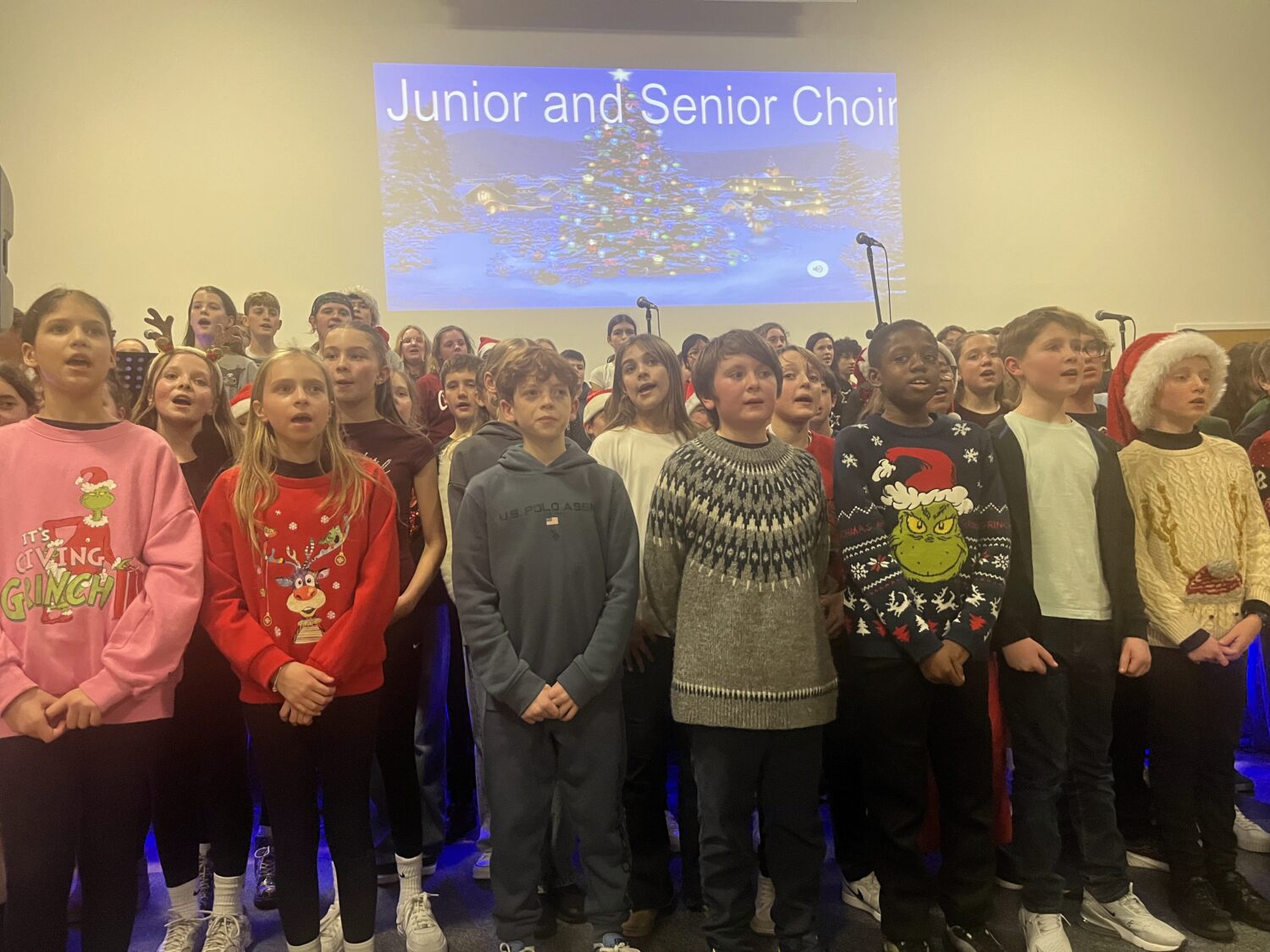 A close-up shot of the "Junior and Senior Choir" featuring students in colorful holiday sweaters and Grinch-themed shirts. They are singing together with focused expressions in front of a festive digital projection.