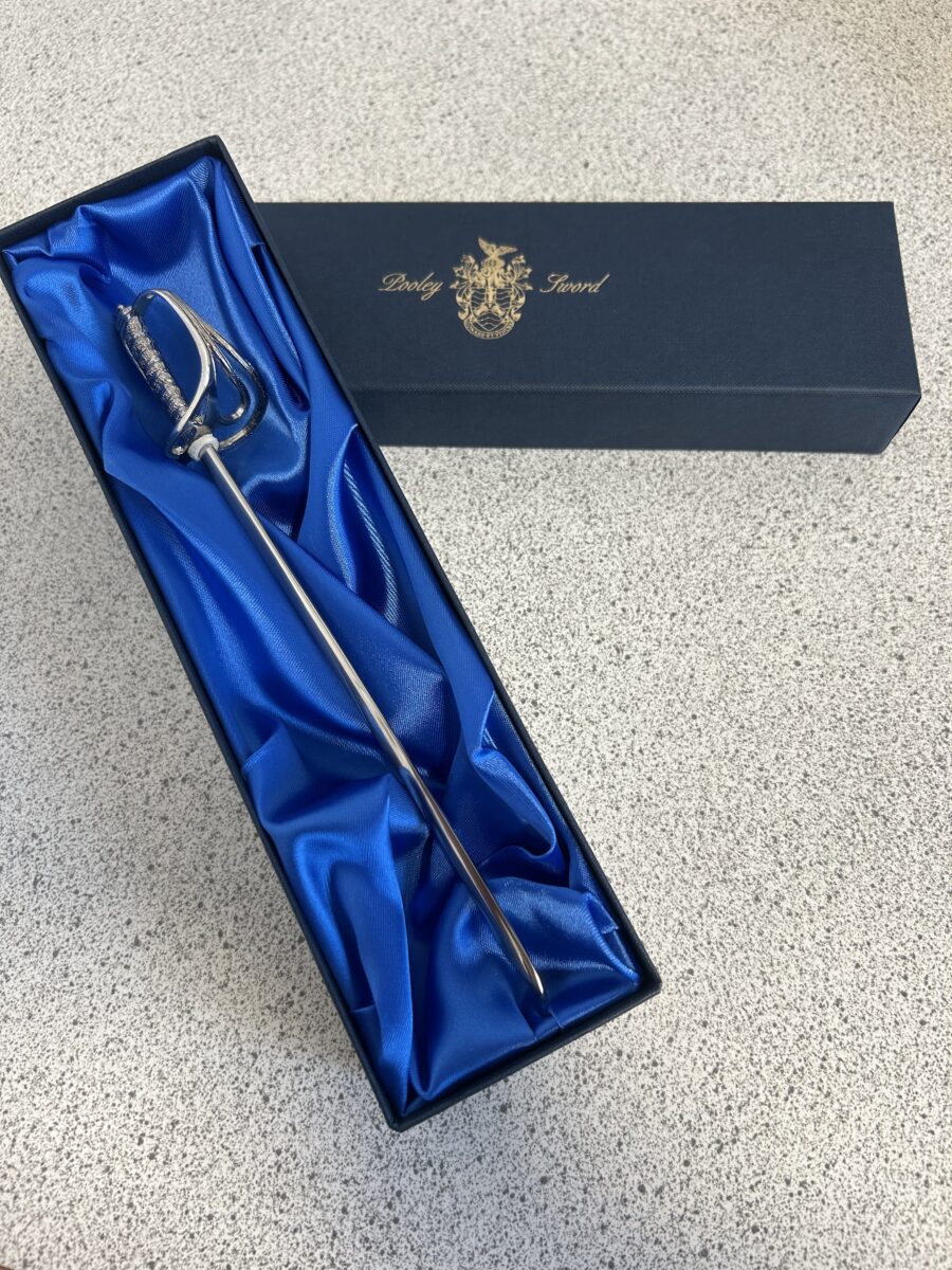 Two individuals, both wearing military camouflage uniforms and black berets, are shaking hands. The older person on the right is presenting a small, dark blue box to the younger person on the left, who is wearing glasses and has a Corporal rank badge.