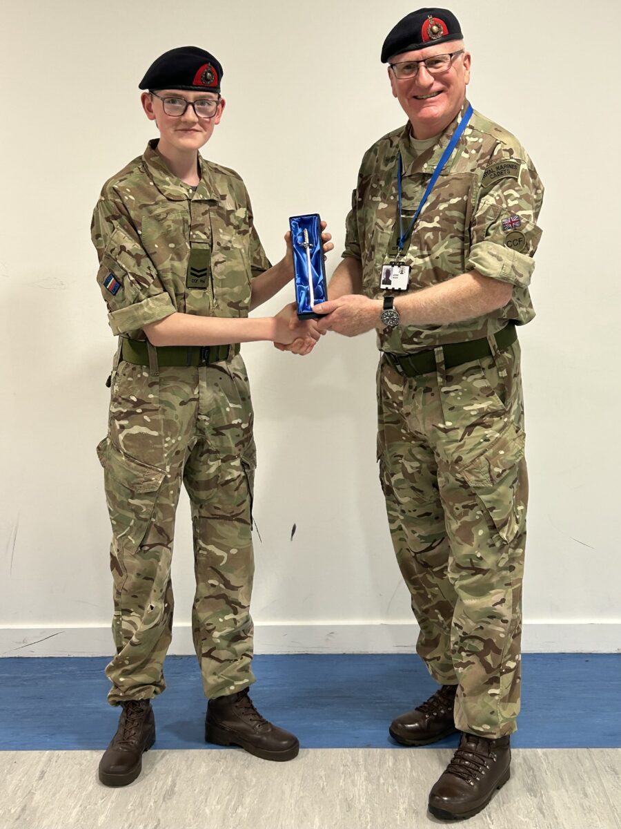 Two individuals, both wearing military camouflage uniforms and black berets, are shaking hands. The older person on the right is presenting a small, dark blue box to the younger person on the left, who is wearing glasses and has a Corporal rank badge.