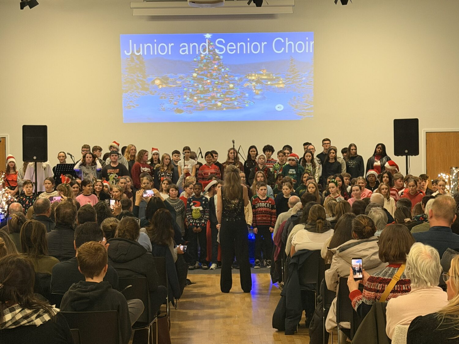 A wide-angle view from the back of a crowded auditorium showing a large student choir on stage. A conductor stands in the center aisle, and several audience members are holding up phones to record the performance.