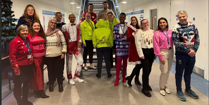 16 people standing in front of stairs wearing Christmas jumpers and outfits.