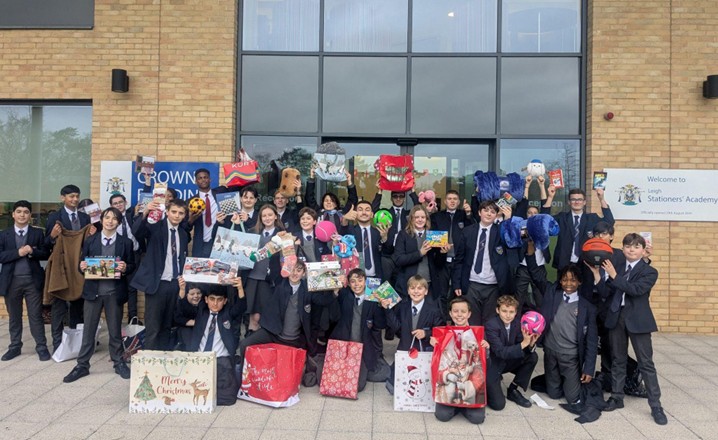 A large group of students in school uniforms stand outside the entrance of "Leigh Stationers' Academy". They are smiling and holding up a variety of holiday gifts, including toys, sports balls, festive gift bags, and books. The sign behind them identifies the building as the Crown Building.