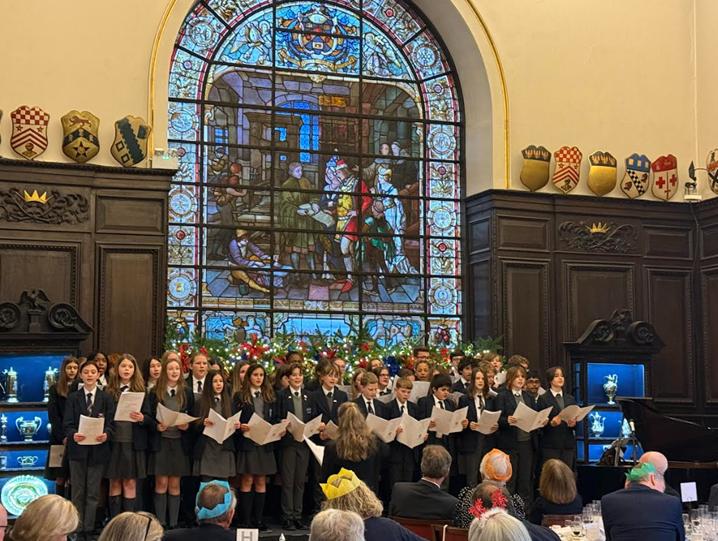 A brief description of the image: A school choir of children in uniform is performing in a grand, wood-paneled hall decorated with formal crests and display cabinets. They are standing directly in front of a large, central stained-glass window with a festive garland underneath. An audience of adults, some wearing Christmas novelty hats, is seated in the foreground watching the performance.