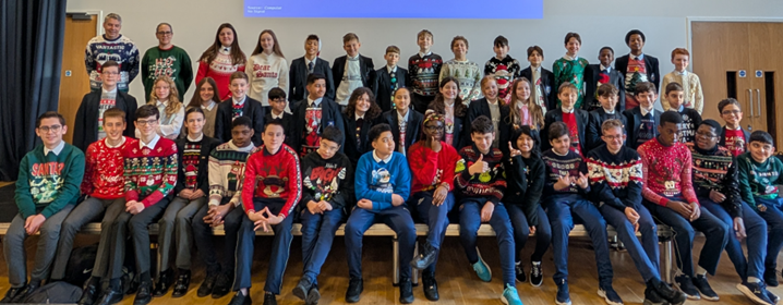 A group of students and a few adults are posing for a photograph indoors. Most of the people, especially the students, are wearing Christmas-themed or "ugly" Christmas sweaters. They are arranged in three rows, with most of the front row sitting down. The background appears to be a school hall or large room with wooden paneling and a large, blank screen above the group.