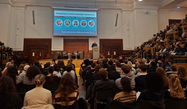 A large audience of students is attending the emagazine A Level Language Conference in a tiered lecture hall. A speaker is presenting from a lectern on the stage, which is dominated by a large screen displaying the conference title, speaker photos, and a reminder for the audience to silence their mobile phones.