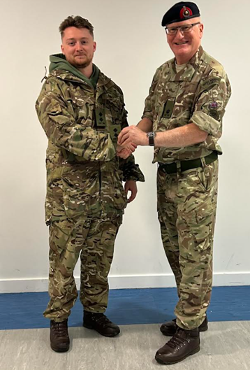 The alt text for this image could be: A full-length indoor photograph showing two men in military camouflage uniform shaking hands. The man on the left is younger, wearing a hooded jacket over his uniform. The man on the right is older, wearing a beret with a military cap badge and glasses, suggesting a higher rank. They are standing in front of a white wall with a blue strip near the floor.