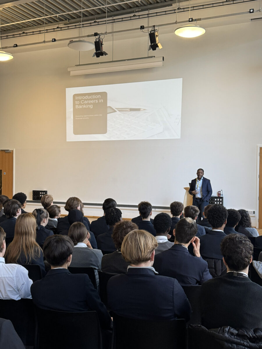 A speaker in a suit stands at the front of a large hall, giving a presentation titled "Introduction to Careers in Banking" to an audience of students seated in rows. The presentation title and the subtitle, "Exploring opportunities within the financial industry," are clearly projected on the wall behind him.