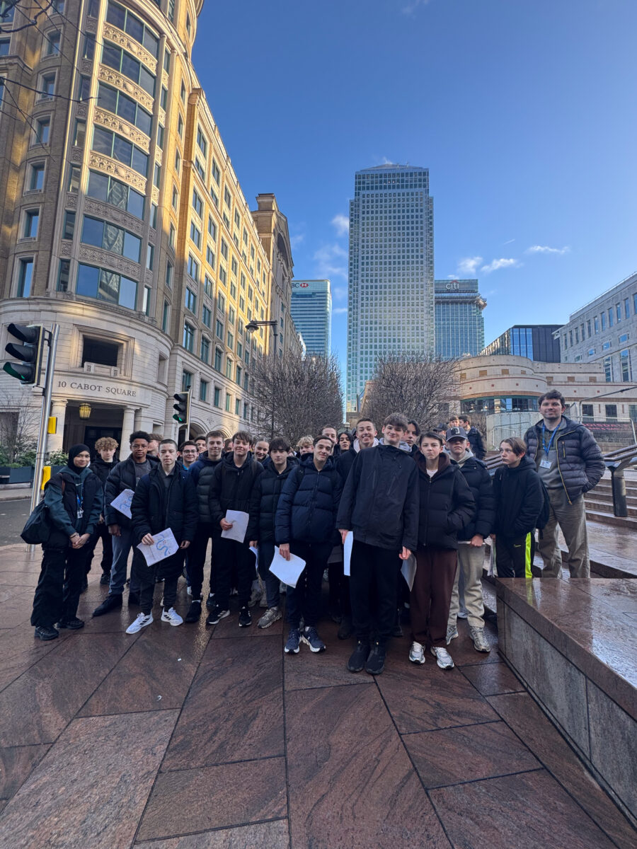 A large group of students stands together in a paved city plaza, with many holding white papers. High-rise office buildings, including towers with HSBC and Citi logos, rise in the background under a clear sky.