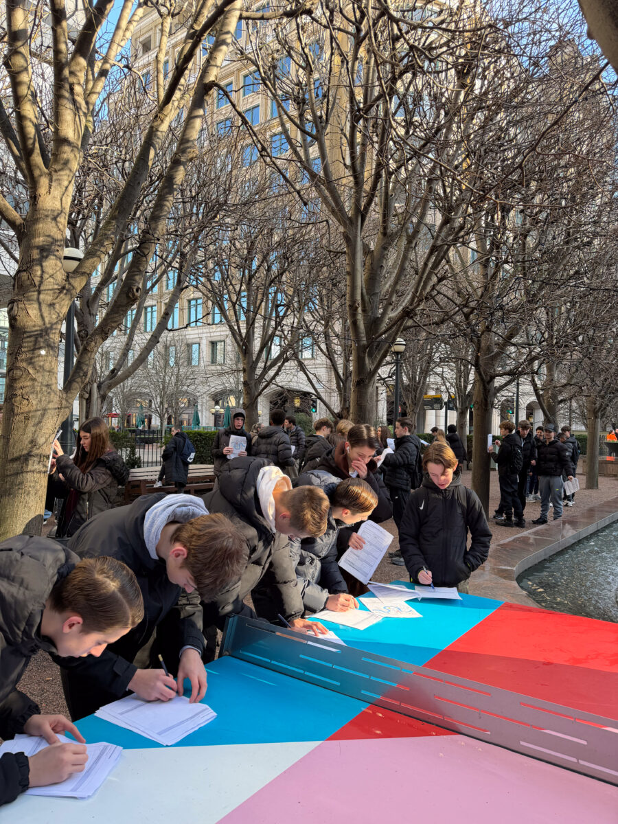 A group of young people lean over a bright blue and red outdoor table to write on papers, while others are scattered among bare trees in the background. The scene takes place in a public urban space featuring modern architecture and a nearby water fountain.