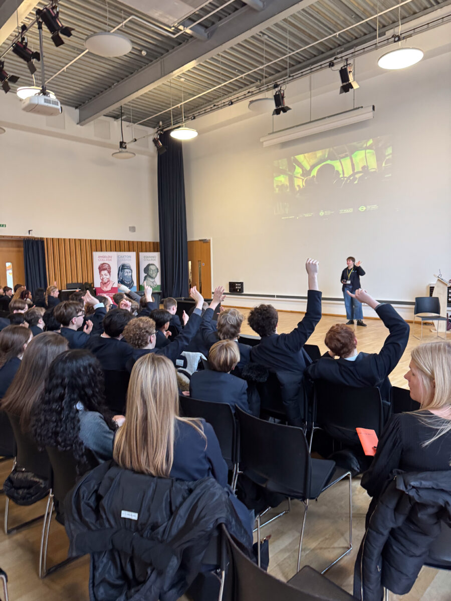 A large group of students, seen from behind, are seated in black chairs in a modern, high-ceilinged assembly hall during a presentation. Several students have their hands raised to participate or ask questions. At the front of the room, a presenter stands before a large projected image on the wall. To the left of the stage area, three banners are visible representing Angelou College, Caxton College, and Equiano College.