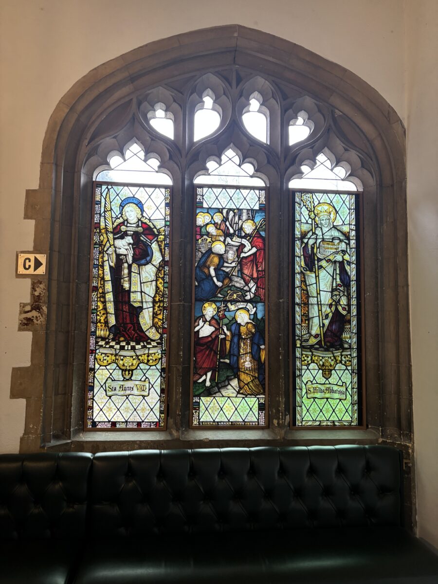A detailed view of a three-panelled stained-glass window set in a stone archway above a dark leather bench.