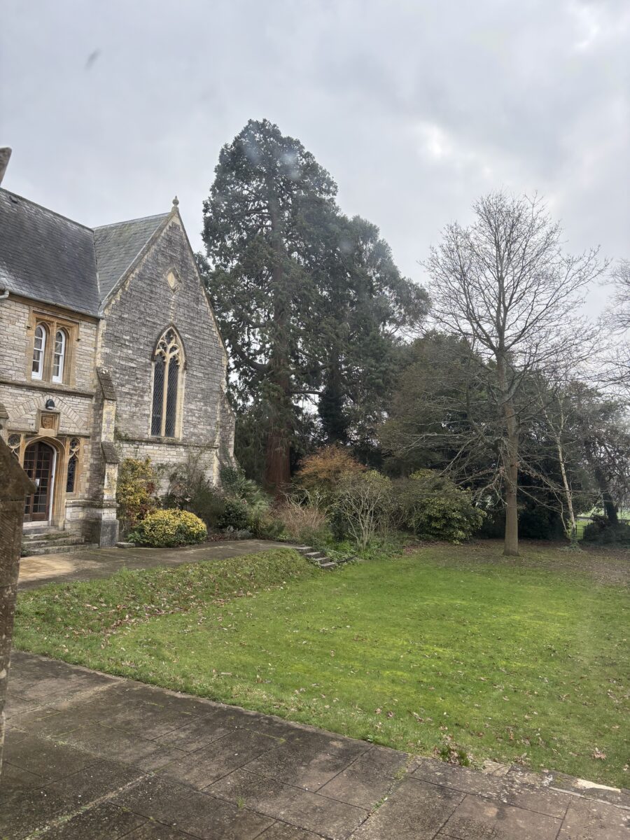 An exterior view of a historic stone building with a large arched window, surrounded by green lawn and tall trees.