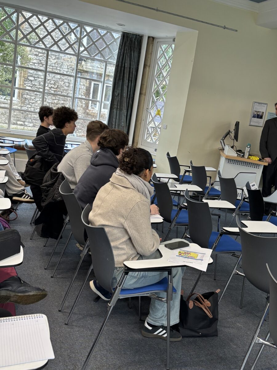 Students seated at desks with blue chairs in a lecture hall, looking toward the front of the room during a presentation.