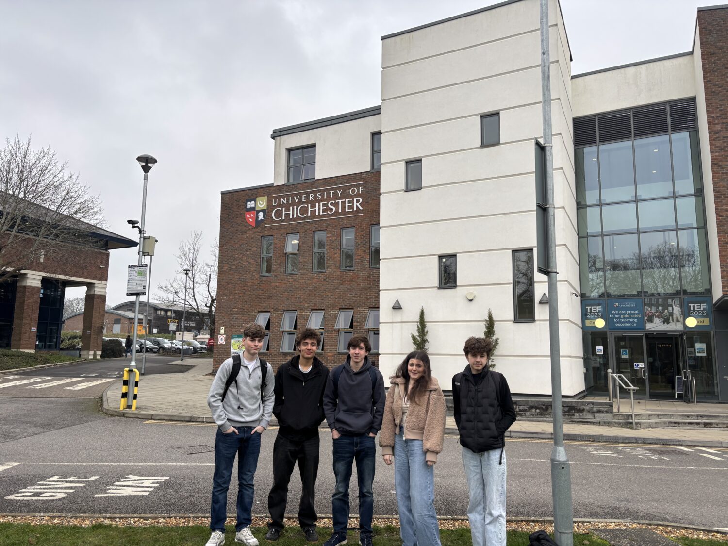 Five students standing in a line outdoors in front of a modern white and brick university building under an overcast sky.