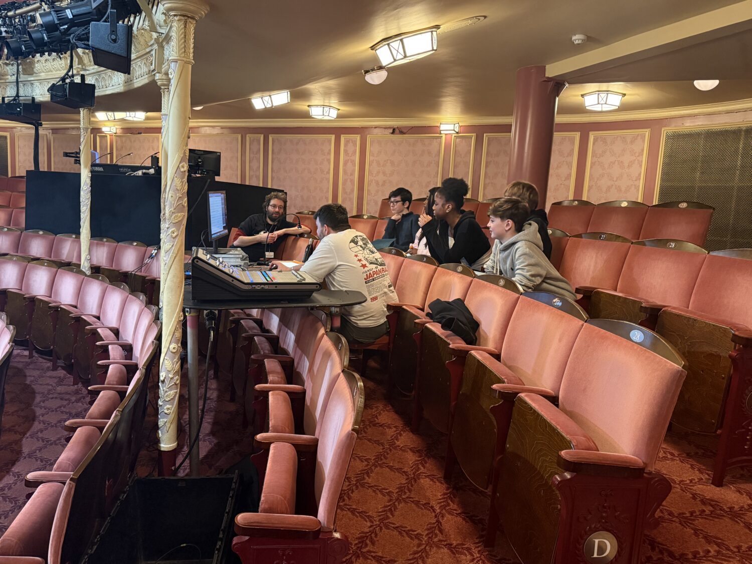 An instructor talks to a group of students seated in rows of theater chairs next to a sound mixing board. The theater features ornate gold pillars and decorative red wallpaper.