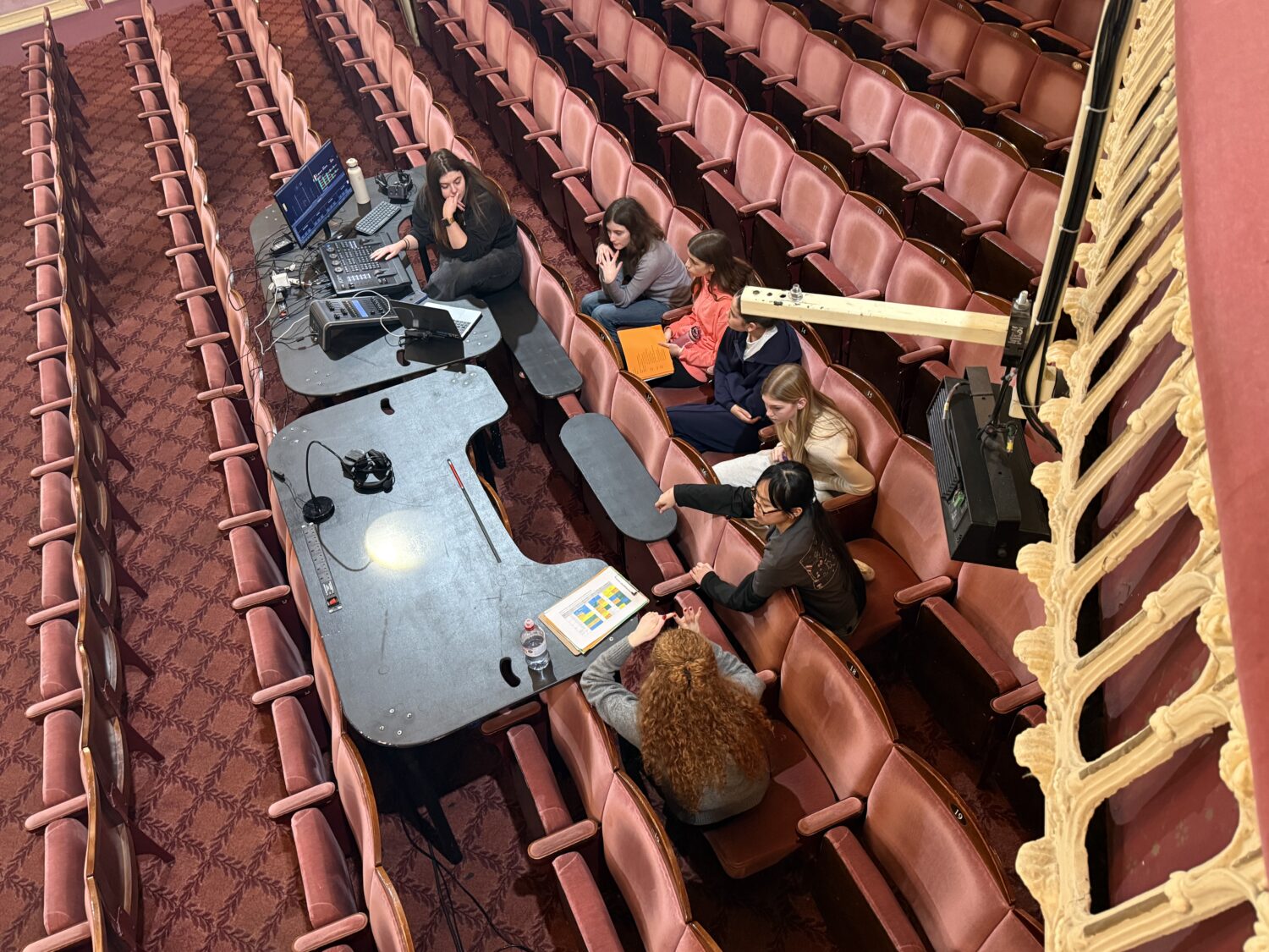 A top-down view of a technical crew working in theater seats. Five people are gathered around a makeshift black desk equipped with audio equipment, laptops, and documents, positioned within the rows of red velvet seating.