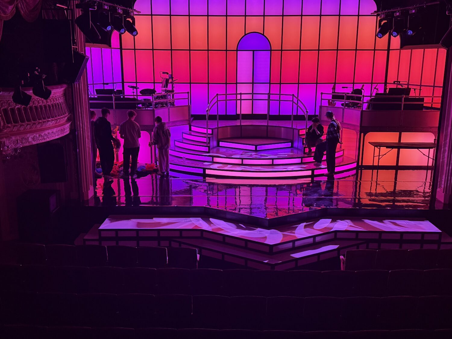 A vibrant theater stage featuring a tiered semi-circular platform and a large grid backdrop illuminated with a pink-to-orange gradient. Several people stand on the reflective black stage floor, which is framed by rows of empty theater seats in the foreground.
