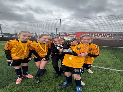 : Five young athletes in yellow and black uniforms pose on a green turf field. Each girl is wearing a gold medal around her neck. One athlete in the center holds up a silver trophy, and a banner with the word "WEMBLEY" is visible in the background under an overcast sky.