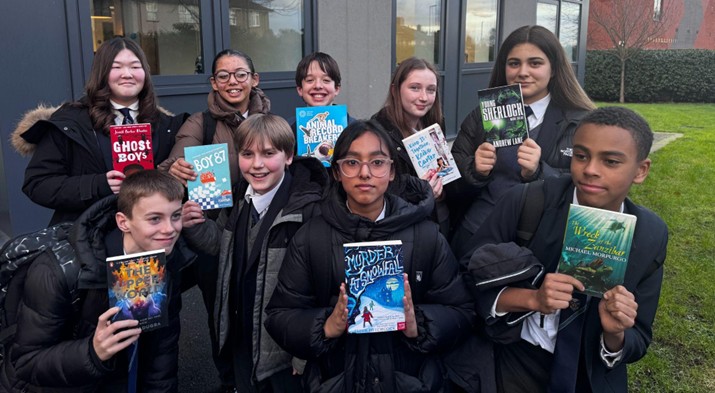 A group of nine students stands outdoors, smiling and holding up various books toward the camera. Some are wearing school uniforms and dark winter coats.
