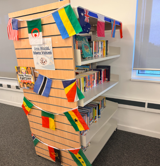 A light-coloured wooden bookshelf end-panel decorated with international flag bunting and a poster that reads "One World, Many Voices."