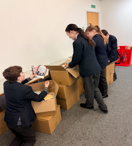 Students in school uniforms kneel and stand around several large cardboard boxes, unpacking stacks of books in a room with white walls.