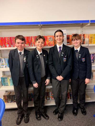 Four students in dark school blazers and grey sweaters stand smiling in front of a white bookshelf filled with colourful books.