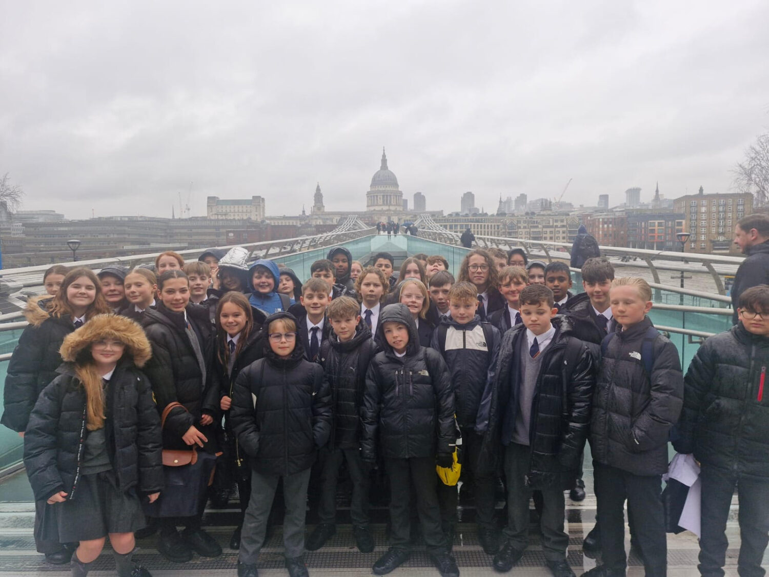 A large group of schoolchildren in dark uniforms and winter coats poses for a group photo on the Millennium Bridge in London. The iconic dome of St. Paul’s Cathedral is centered in the background under a grey, overcast sky. The river and the city skyline are visible on either side of the bridge.