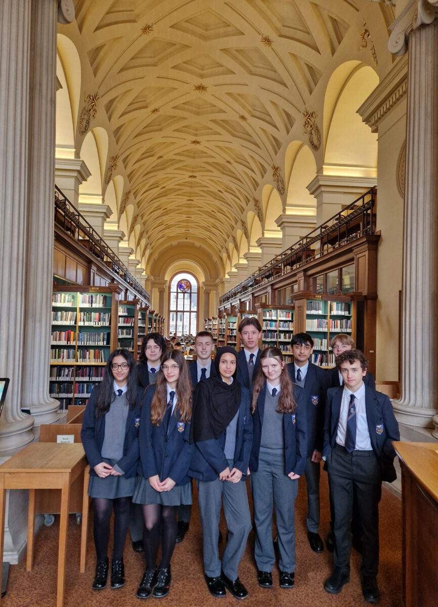 Ten students pose together in a grand, historic library with a very high, ornate vaulted ceiling. They are framed by massive stone columns and long rows of tall, dark wood bookshelves.