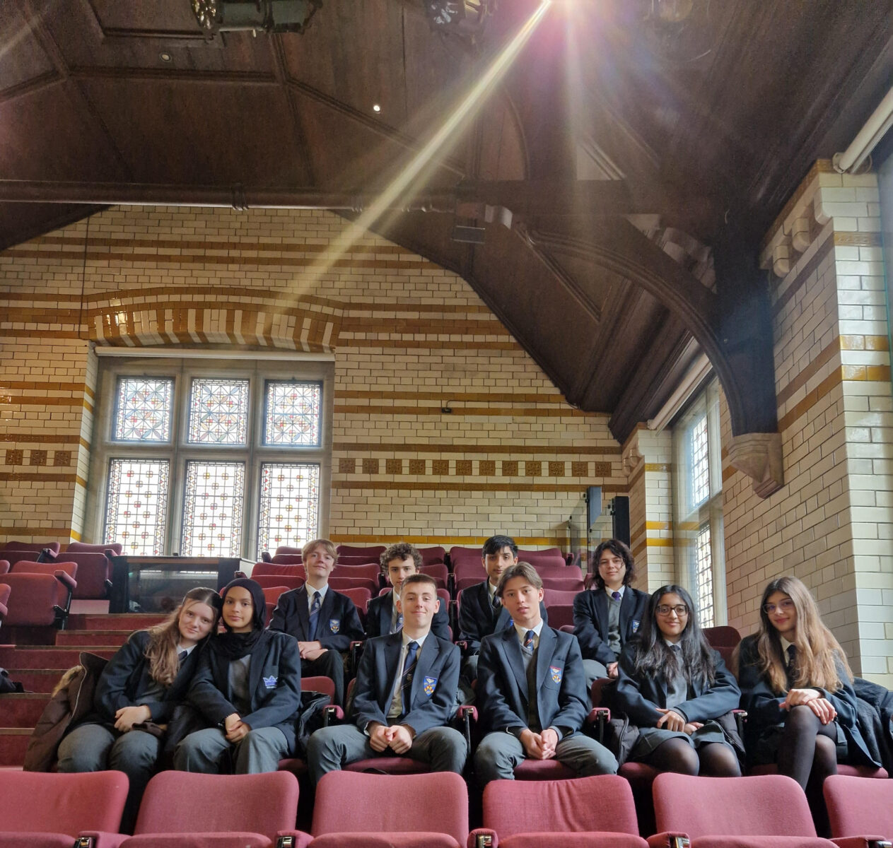 A group of ten students in matching school blazers sits in tiered red theatre seating. The room features high, vaulted wooden ceilings and large stained-glass windows set into patterned brick walls.