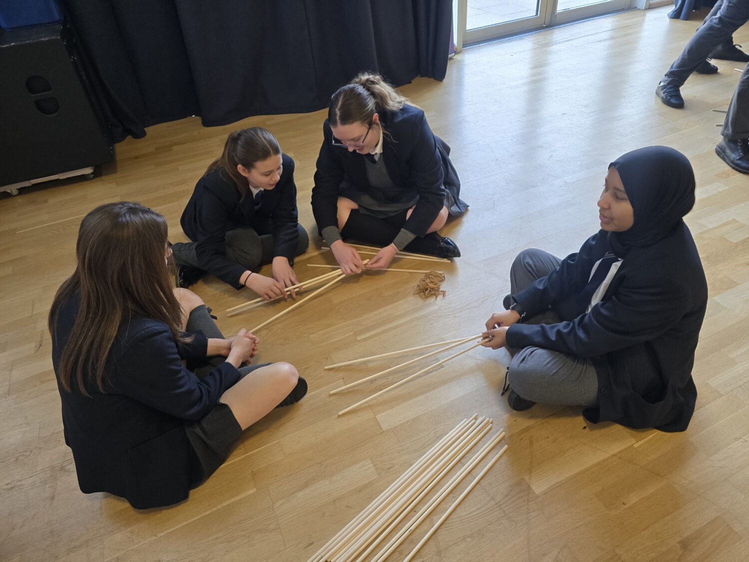 Four students sit in a circle on a wooden floor, working together to connect long wooden dowels using rubber bands to form the base of a structure.