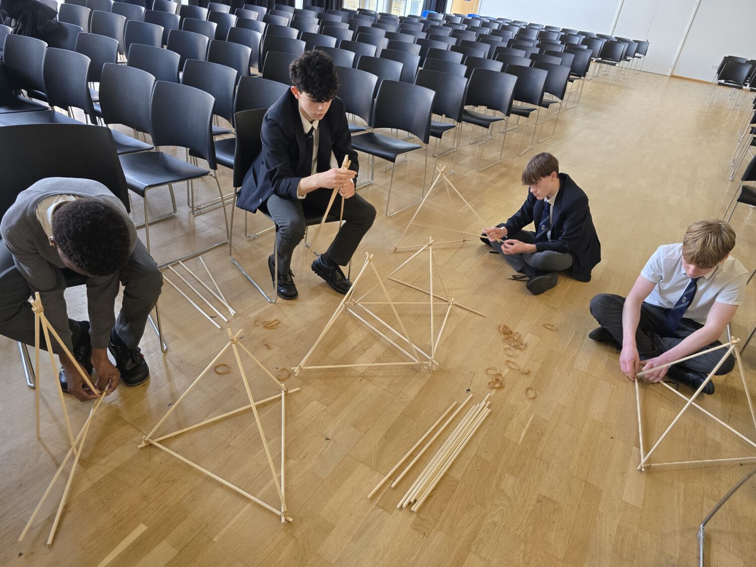 Several students sit on the floor or chairs in a large hall, each working individually on constructing small, three-dimensional triangular frames (tetrahedrons) from wooden sticks.