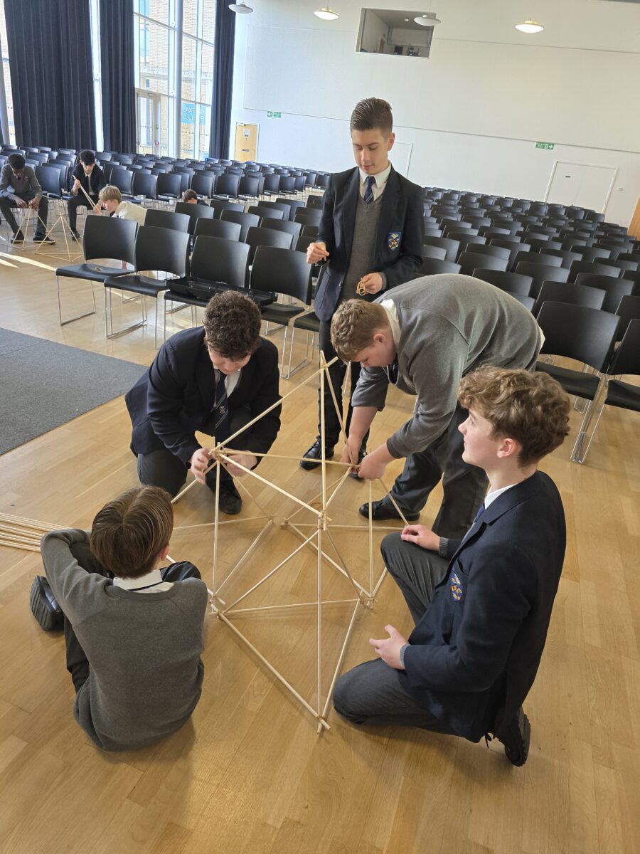 A group of four students in school uniforms huddles together on the floor, carefully attaching a new wooden dowel to a growing geometric structure.